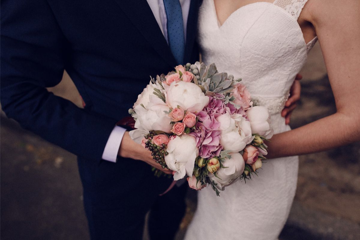 On their wedding day, a bride in a white dress holds a bouquet of pink and white flowers, standing beside a groom in a dark suit and blue tie. Their heads are out of shot.