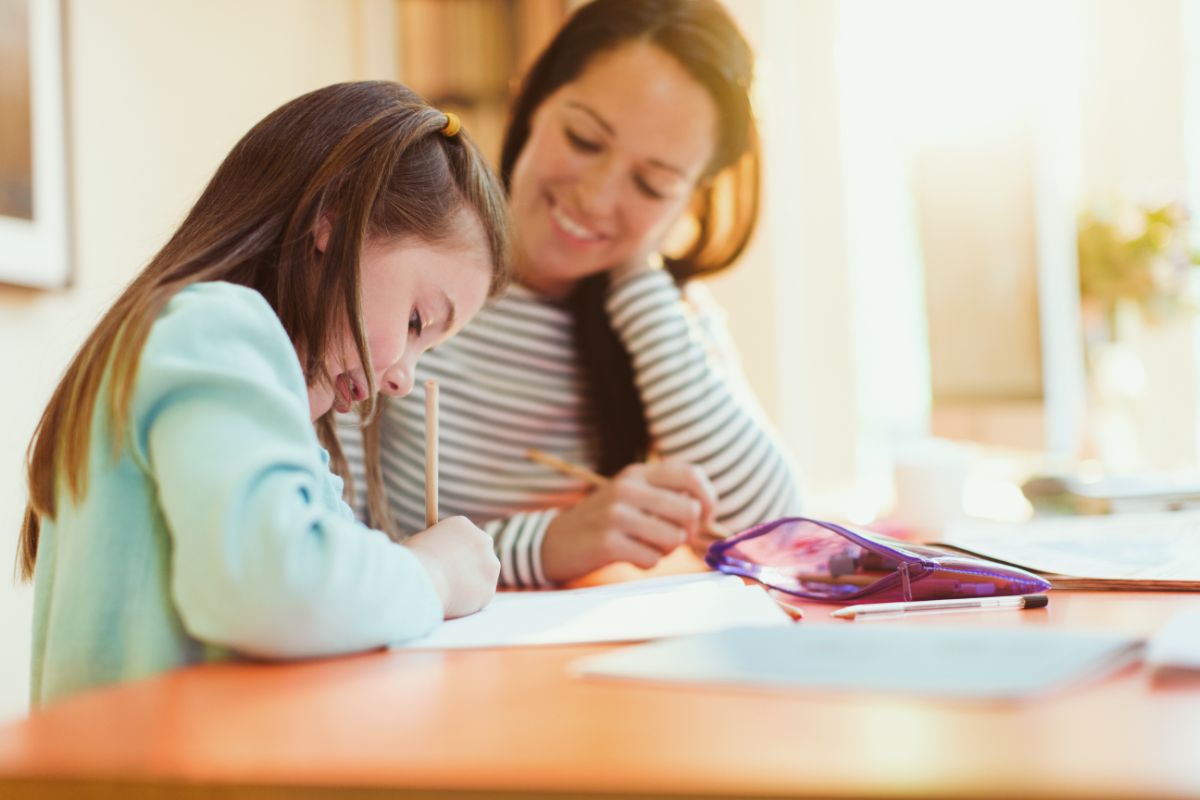 A young girl writes in a notebook at a table during an after-school session whilst an adult woman, possibly her mother or teacher, sits beside her smiling. Sunlight fills the bright, cosy room. A pencil case and paper are on the table.
