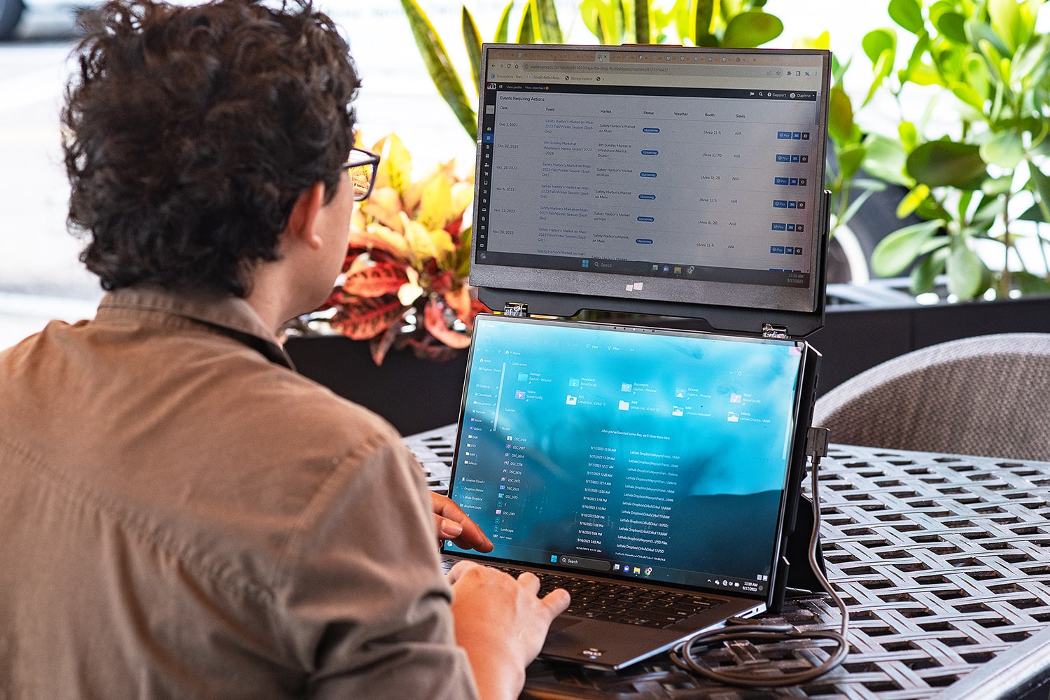 A person with dark hair works on a laptop with an extra screen attached above it, both displaying different content, while sitting at an outdoor table surrounded by plants and enjoying a perfect ergonomic set-up.