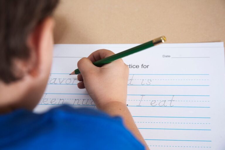 A child practises handwriting with a green pencil on lined paper, tracing cursive letters. Wearing a blue shirt, only the back of their head and hand can be seen as they focus on improving their handwriting skills.