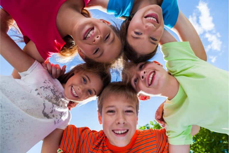 Five smiling children stand in a circle outdoors, heads together and looking down at the camera, each radiating happiness as they celebrate one another's child's strengths against a bright blue sky and lush greenery.