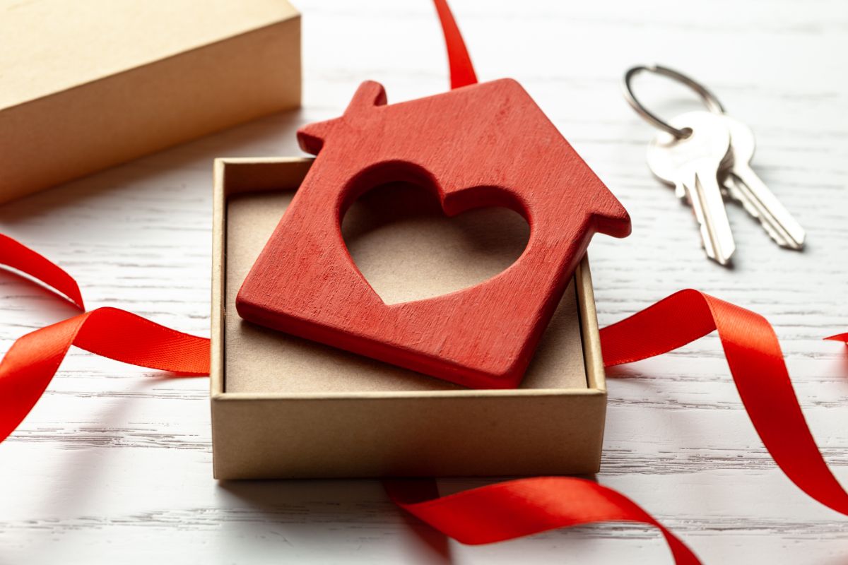 A small red wooden house with a heart cut-out sits in an open gift box next to a red ribbon, with two keys on a keyring nearby—a perfect housewarming present—displayed on a white wooden surface.