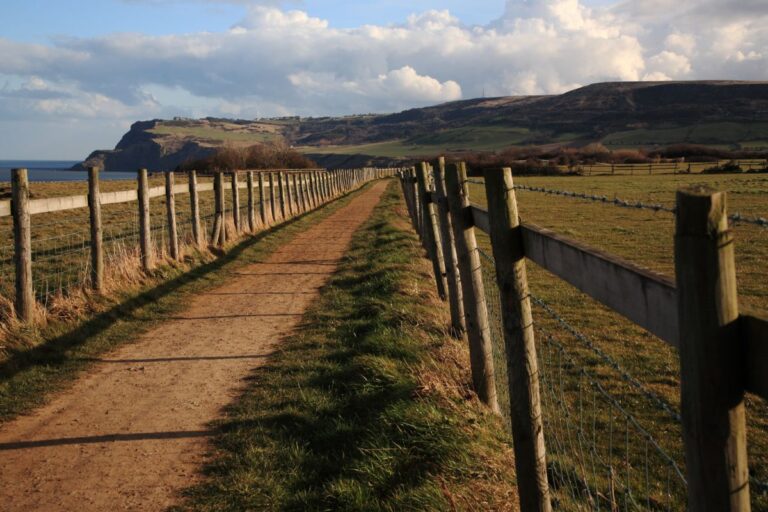 A dirt track lined with wooden fences runs through grassy fields towards distant hills, with the sea visible on the left and a partly cloudy sky overhead—perfect for relaxed family days out.