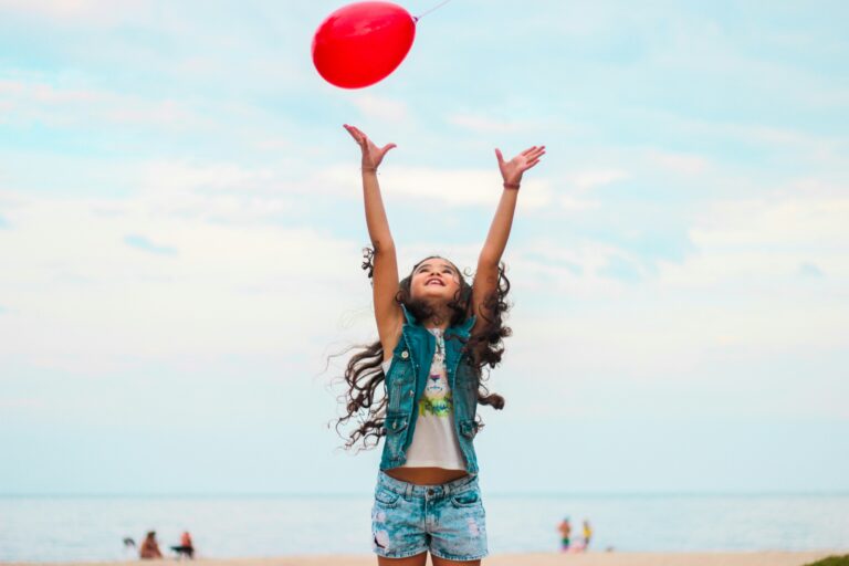 A young girl with long curly hair and denim clothes confidently reaches up to release a red balloon on a sandy beach, with people and the sea in the background under a cloudy sky.