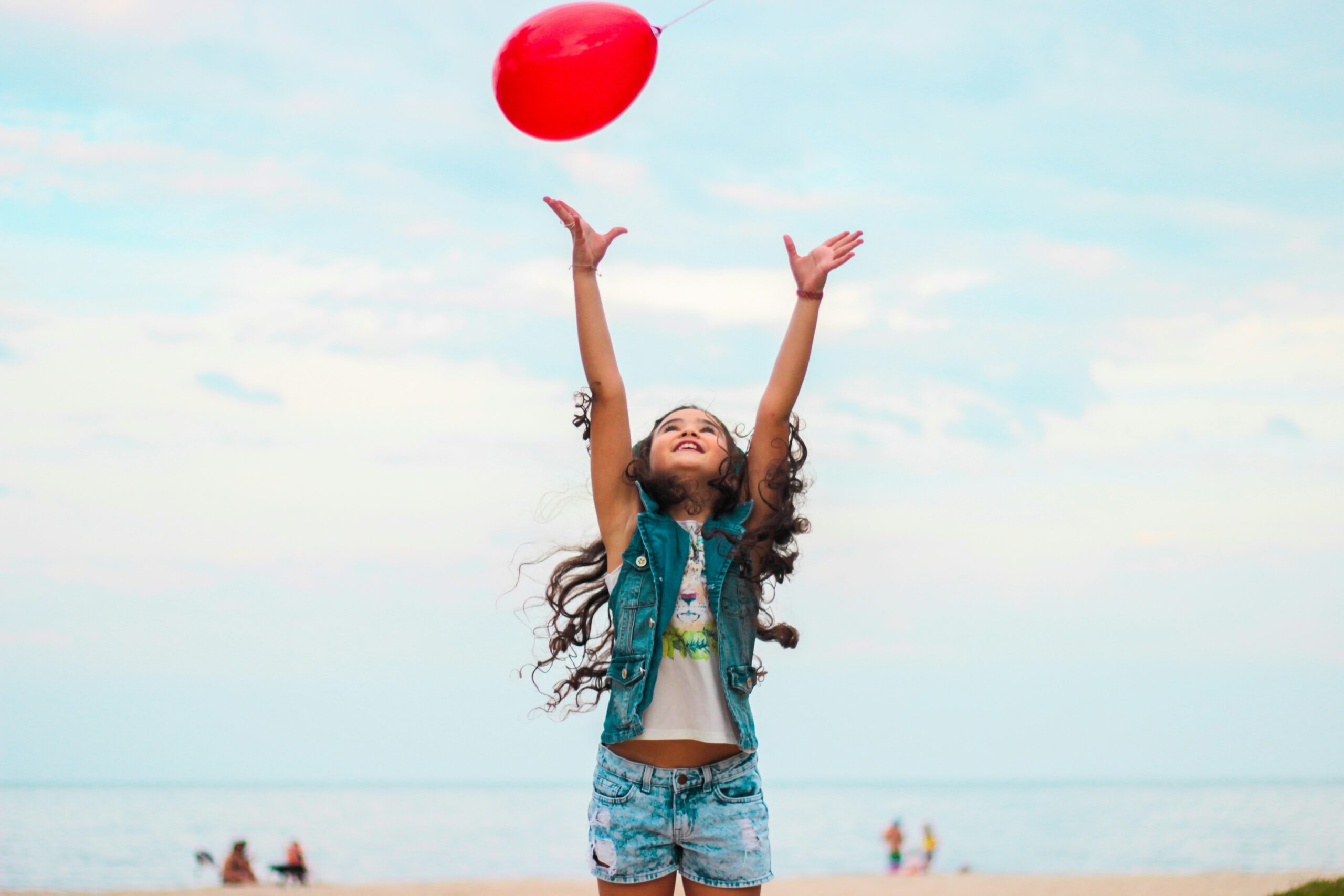 A young girl with long curly hair and denim clothes confidently reaches up to release a red balloon on a sandy beach, with people and the sea in the background under a cloudy sky.