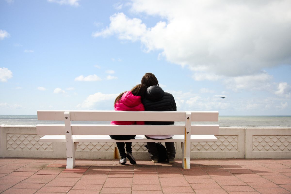 Three people wearing jackets sit closely together on a white bench, possibly sharing a quiet dating moment as they face the sea. With their backs to the camera and a partly cloudy sky above, the scene feels peaceful and serene.