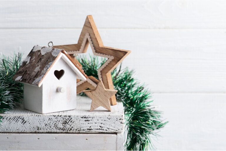 A small white birdhouse with a heart-shaped cut-out sits next to two wooden star decorations on a rustic white surface, creating a cosy scene of a home ready for Christmas. Green garland decorates the background against a white wooden wall.