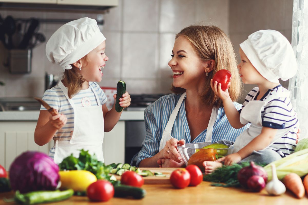 A smiling woman and two young children wearing chef hats sit in a kitchen, surrounded by fresh vegetables. As they cook together, laughing and holding veg, they show how fun it can be to eat healthily as a family.