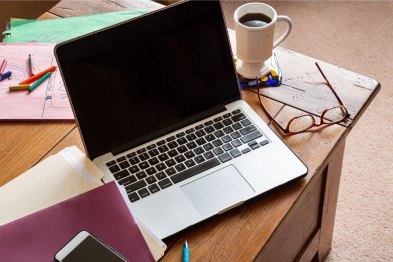 A laptop on a wooden table surrounded by papers, a mobile phone, coloured pens, spectacles, and a coffee mug—a busy, informal workspace reflecting the dynamic life of a mum in business.