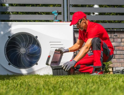 A technician in red uniform kneels on grass, opening a toolbox near an outdoor heat pump unit, with a brick wall and metal fence in the background.