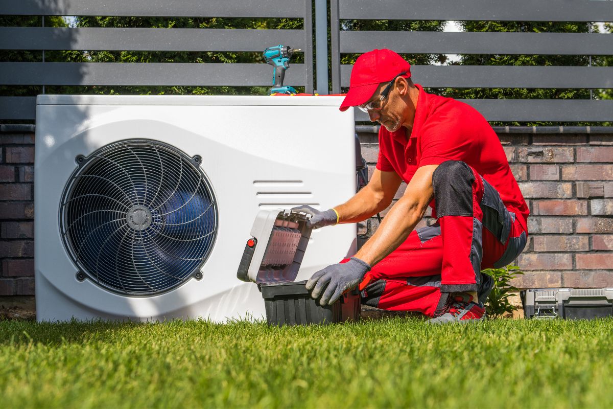 A technician in red uniform kneels on grass, opening a toolbox near an outdoor heat pump unit, with a brick wall and metal fence in the background.