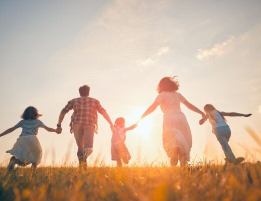 A family of five is holding hands and running through a grassy field at sunset, the sunlight casting a warm, glowing atmosphere—capturing the joy and chaos of life with three children.