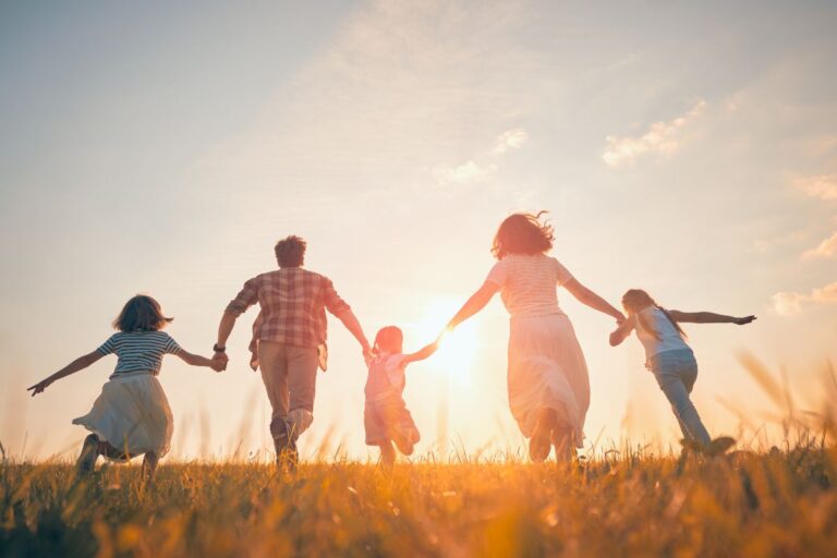 A family of five is holding hands and running through a grassy field at sunset, the sunlight casting a warm, glowing atmosphere—capturing the joy and chaos of life with three children.