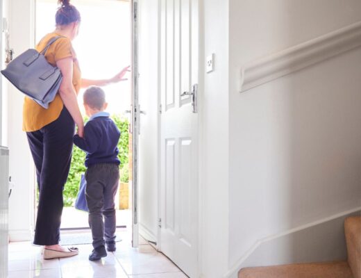 A woman holding a young child's hand stands near an open front door, ready for the school run. She carries a blue bag as sunlight streams in, illuminating the hall and staircase—capturing the everyday moments parents survive with helpful parenting tips.