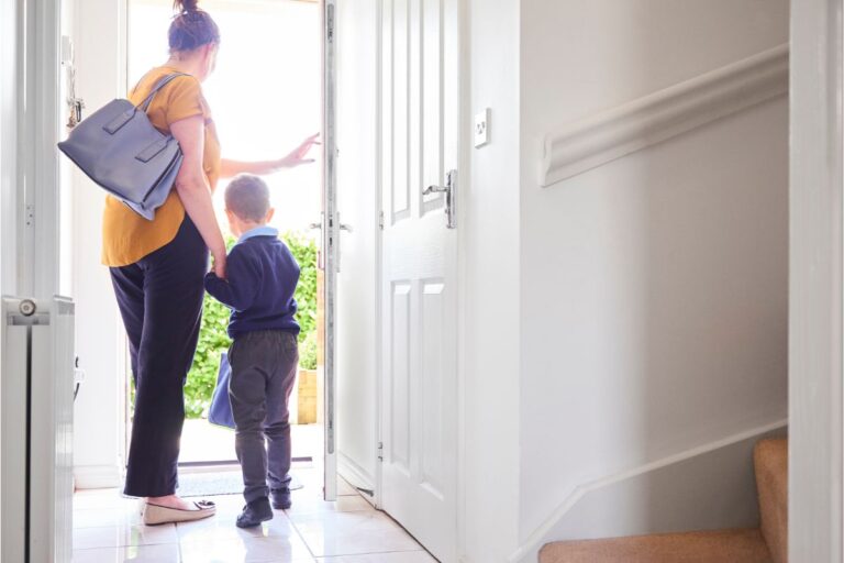 A woman holding a young child's hand stands near an open front door, ready for the school run. She carries a blue bag as sunlight streams in, illuminating the hall and staircase—capturing the everyday moments parents survive with helpful parenting tips.