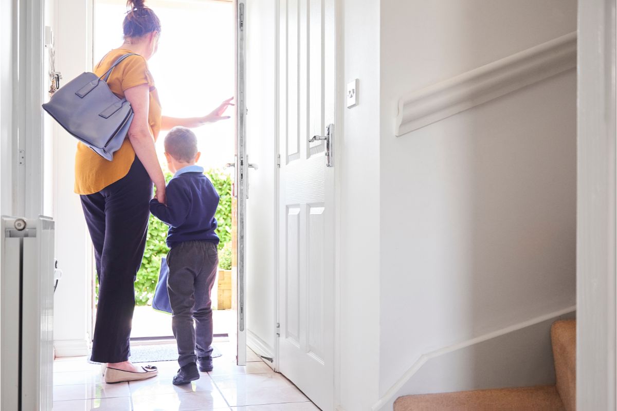 A woman holding a young child's hand stands near an open front door, ready for the school run. She carries a blue bag as sunlight streams in, illuminating the hall and staircase—capturing the everyday moments parents survive with helpful parenting tips.