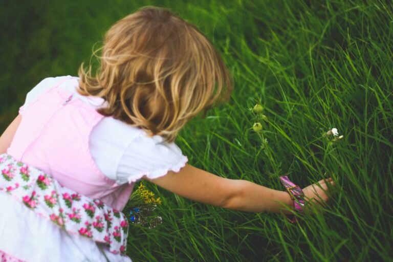 A young child wearing a pink dress with floral patterns is kneeling in tall green grass, reaching out to pick small flowers with one hand. The scene suggests a peaceful, outdoor setting.