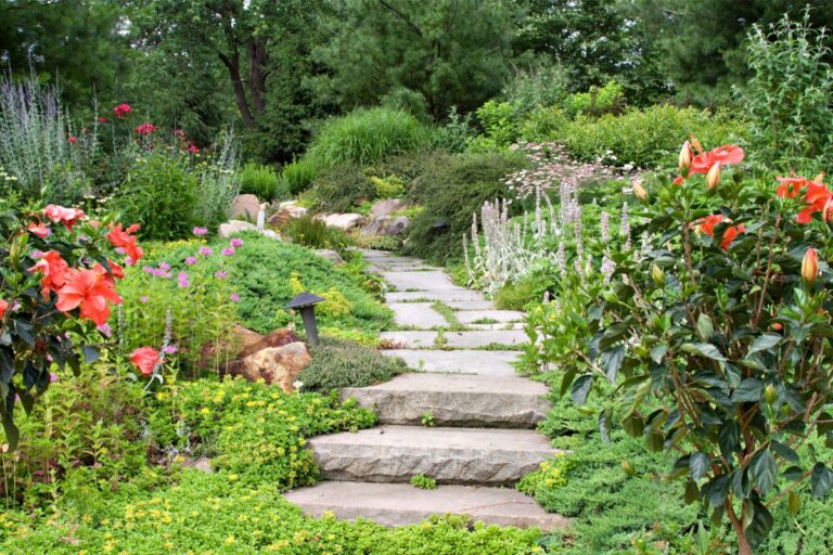 A stone path with steps winds through a lush garden filled with colourful flowers, green shrubs, and tall trees. A small garden light sits beside the path—perfect inspiration for budget garden hacks to create beauty without overspending.