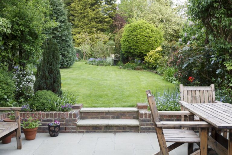A neatly mown lawn bordered by lush trees, shrubs, and flowerbeds, viewed from a stone patio with wooden outdoor furniture and potted plants. Stone steps connect the patio to the garden.