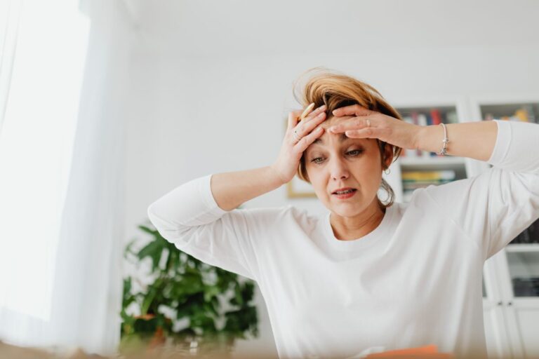 A woman in a white shirt sits indoors, holding her head with both hands and looking stressed—capturing the invisible mental load that mums often face. There is a blurry plant and bookcase in the bright background.