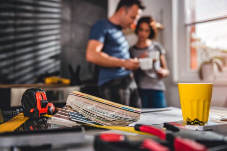 A tape measure, colour swatches, and tools are on a table in focus—essential items for any homebuilding project. In the background, a man and woman stand together at what could be the Harrogate Renovating Show, looking at a piece of paper, with a yellow mug and sunlight streaming through the window.